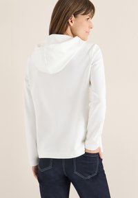 Sudadera blanca de corte relajado, que incluye una capucha con cordón y mangas largas. Combinada con unos jeans oscuros, mostrando un atuendo casual.