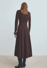 Robe marron à manches longues avec une jupe évasée, en tissu lisse. Associée à de longues bottes noires et des bracelets en bois au poignet.