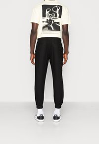 Zwarte joggingbroek met elastische boorden, gecombineerd met een crèmekleurig T-shirt met een groot zwart grafisch ontwerp op de achterkant. Witte sokken en sneakers.