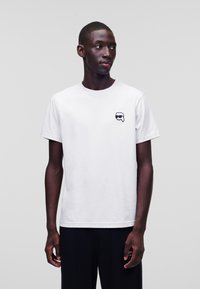 Witte katoenen t-shirt met ronde hals, korte mouwen en een klein zwart grafisch logo op de linkerkant van de borst. Model draagt zwarte broeken.