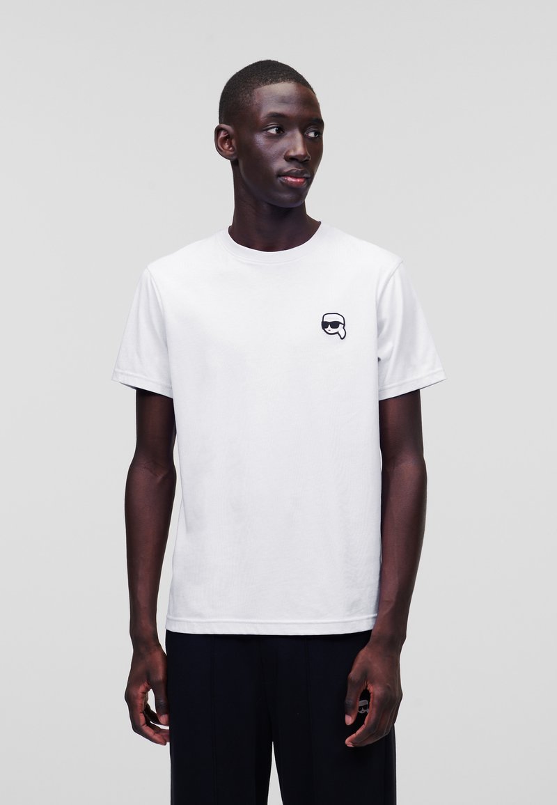 Witte katoenen t-shirt met ronde hals, korte mouwen en een klein zwart grafisch logo op de linkerkant van de borst. Model draagt zwarte broeken.