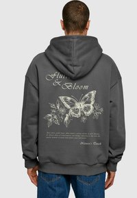 Mørkegrå hættetrøje med et stort hvidt sommerfugle- og blomsterdesign på ryggen, samt dekorativ tekst nedenfor.