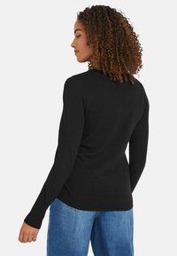 Frau mit lockigem Haar, die ein enges schwarzes Langarmoberteil und eine blaue Jeans trägt, nach links blickend, wobei ihr Rücken größtenteils sichtbar ist.