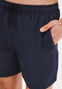 Granatowe kąpielówki z elastycznym pasem, regulowanym sznurkiem, bocznymi kieszeniami i gładką fakturą, z minimalistycznym logo na brzegu.