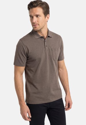 Mann trägt ein braunes kurzärmeliges Poloshirt mit Brusttasche, steht und blickt nach links vor einem einfarbigen weißen Hintergrund.