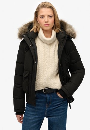 Sort pufferjakke med pelsforet hætte, forlommer og ribbede manchetter. Båret over en cremefarvet kabelstrikket sweater og blå jeans.