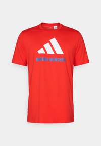 Rød bomull t-skjorte med en hvit tre-stripes logo og "BAYERN MÜNCHEN" i blått. Rund hals og korte ermer, enkelt design.