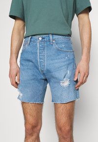 Blue denim shorts featuring a frayed hem, distressed patches, and classic five-pocket design. Worn with a green t-shirt.