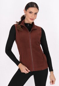 Vesta din fleece maron, cu fermoar în față, guler înalt și buzunare laterale. Puritată peste o cămașă neagră cu mâneci lungi, având o textură fină.