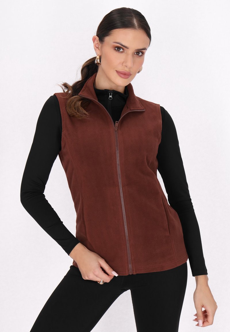 Chaleco de forro polar marrón con cremallera frontal, cuello alto y bolsillos laterales. Lleva puesto un camiseta de manga larga negra, con una textura suave.