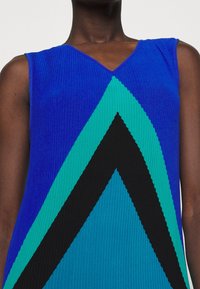 Haut sans manches côtelé bleu avec un col en V, présentant des motifs géométriques audacieux en turquoise et noir, créant un contraste visuel saisissant.