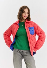 Žena v červené prošívané bundě s modrým kapsičkou přes zelený svetr a šedé džíny, pózující se založenýma rukama v pase na bílém pozadí.