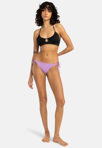 Black bikini top with a front cut-out, paired with light purple tie-side bottoms. Smooth fabric, minimalist design, and adjustable straps.