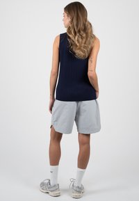Femme aux longs cheveux ondulés portant un top sans manches bleu marine, un short gris clair à hauteur des genoux, des chaussettes blanches et des baskets grises, debout, de dos.