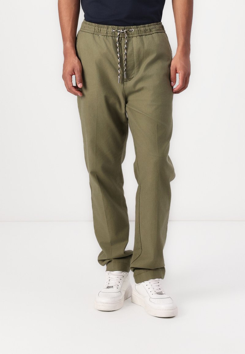 Olivgrüne Jogginghosen mit einem elastischen Bund, silbernem Kordelzug, seitlichen Taschen und einem schmal zulaufenden Bein-Design. Kombiniert mit weißen Sneakers.