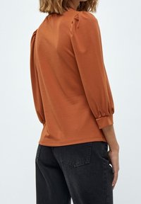 Kovově červený dlouhý rukáv s nabíranými balónovými rukávy a kulatým výstřihem, vyrobený z hladké látky. V kombinaci s tmavými džíny.