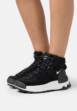 Baskets montantes Nike noires et blanches avec semelles épaisses portées sur des jambes nues, vues sous un angle latéral avant sur fond blanc.