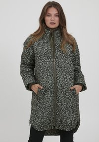 Zaļa quilted jaka ar leoparda rakstu, augstu apkakli un rāvējslēdzēja aizdari. Iespējamas sānu kabatas un garāks apakšmalas garums aizmugurē.