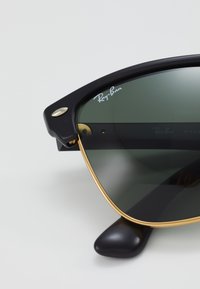 Ochelari de soare Ray-Ban negri cu lentile verzi, accente de metal auriu și un cadru texturat negru. Logo-ul bine cunoscut afisat pe lentila exterioară.