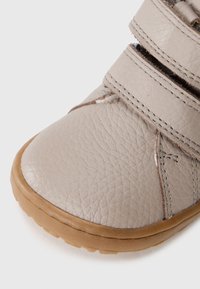 Beige leren kinderschoen met textuur, ronde neus, bruine rubberen zool en twee klittenbandsluitingen.