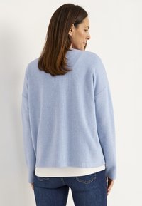 Vaaleansininen raidoitettu neulepaita pitkähihaisilla hihoilla, rento malli ja hieman pyöristetty helma, yhdistettynä tummansinisiin farkkuihin. Pehmeä rakenne on näkyvissä.