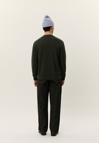 Άνδρας που φοράει γκρι σκουφί, σκούρο πράσινο πουλόβερ, μαύρο τζιν και μαύρα παπούτσια, στέκεται μπροστά σε ένα απλό ανοιχτό фон.