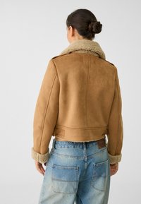 Brązowa krótka kurtka z zamszu z puszystym beżowym kołnierzem i mankietami. Charakteryzuje się nowoczesnym krojem i widocznymi detalami przeszyć. Noszona z niebieskimi dżinsami.