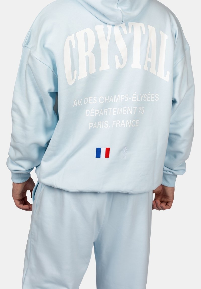 Survêtement bleu clair avec le texte « CRYSTAL », adresse à Paris et petit drapeau français dans le dos, porté par une personne debout de dos.
