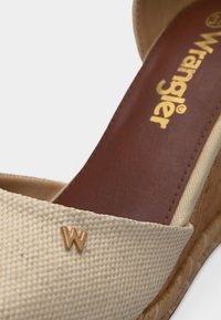 Κοντινό πλάνο μπεζ πλεκτού σανδάλιου με τακούνι φελού σε σφήνα, με χρυσό λογότυπο "W" στο λουράκι και καφέ πάτο με χρυσή σήμανση Wrangler.