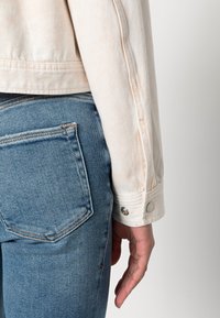 Ljusbeige denimjacka med långa ärmar och knäppningsmanschetter. Bärs tillsammans med ljusblå jeans som har en bakficka och en strukturerad tyg.
