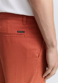 Pantalons en terracotta fabriqués dans un tissu léger, dotés d'une poche arrière avec une petite étiquette noire. Texture lisse et coupe droite.