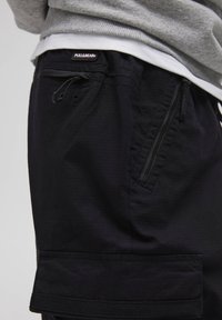 Czarne szorty cargo z teksturowanej tkaniny, z boczną kieszenią na zamek błyskawiczny i paskiem z regulacją na sznurki. Widoczna naszywka z logo na pasie.