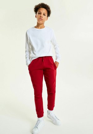 Hvid langærmet skjorte og røde sweatpants med elastisk talje og lommer. Model iført hvide sneakers, stående foran en neutral baggrund.