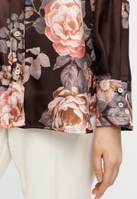 Καφέ μπλούζα με λουλουδάτο μοτίβο που παρουσιάζει μεγάλα ροζ και μπεζ τριαντάφυλλα, με απαλή σατέν υφή, λεπτομέρειες με κουμπιά στις μανσέτες και ελαφρώς καμπυλωτό τελείωμα.