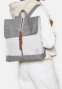 Szary i biały plecak z materiału z klapą, ze szczegółami ze skóry i logo Herschel. Posiada górny uchwyt oraz regulowane paski.