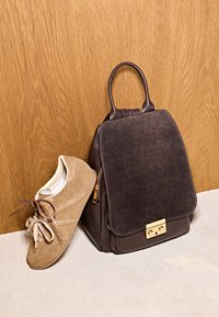 Baskets en daim marron appuyées contre un sac à dos en cuir et daim marron foncé avec une fermeture dorée, posés sur un sol beige devant un mur en bois.