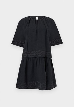 Jonge vrouw staat tegen een effen muur, draagt een zwarte top met korte mouwen en een rok, houdt een strooien handtas met franjes vast en draagt bruine sandalen.