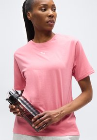 Γυναίκα με ροζ μπλούζα Puma κρατάει ένα μαύρο και διάφανο μπουκάλι νερού, κοιτάζοντας αριστερά της μπροστά σε λευκό φόντο.