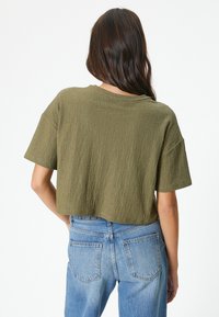 Top corto de manga corta en tejido texturizado verde oliva. Combinado con jeans azules de tiro alto, que muestran un ajuste relajado y un estilo casual.