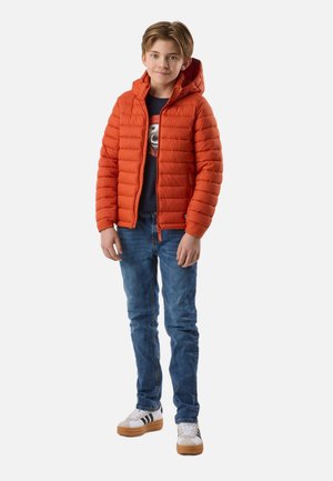 Junge trägt eine orangefarbene Steppjacke, blaue Jeans und weiße Turnschuhe, steht mit entspannten Händen seitlich am Körper.
