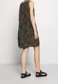 Person klädd i en ärmlös svart och brun klänning med geometrisk mönstring och svarta remsandaler, stående mot en vit bakgrund.