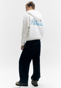 Witte hoodie met blauwe grafische tekst op de achterkant, gecombineerd met donker denim cargobroek en beige schoeisel. Heeft een losse pasvorm en een hoodie met trekkoord.