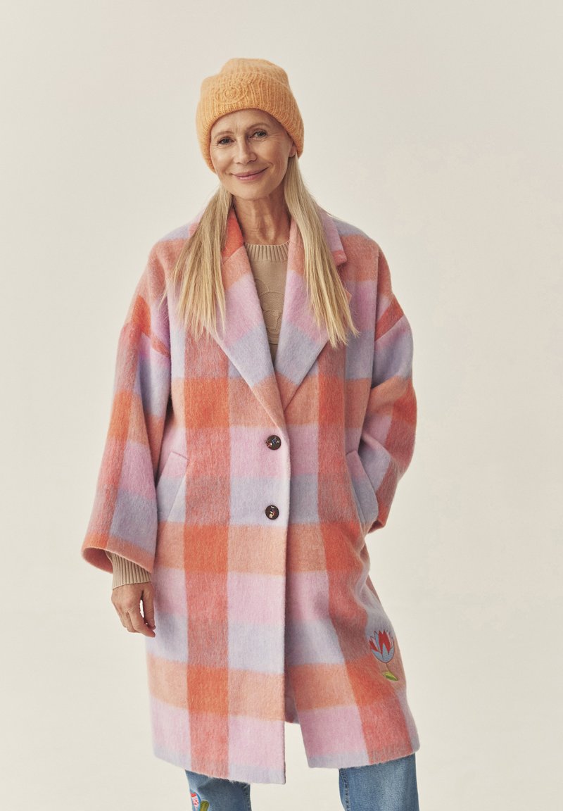 Ülerahvastatud roosa ja oranži ruuduline villane mantel, millel on suured nööbid, plaastritaskud ja lilleline tikanddetail, kantud beeži kampsuni ja teksade peale.