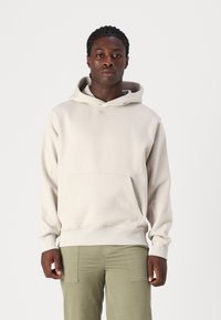 Light beige hoodie with a front kangaroo pocket, ribbed cuffs, and a drawstring hood, made from soft cotton fabric. Worn over olive green pants.