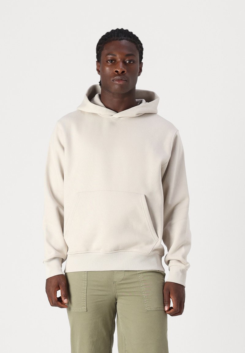 Light beige hoodie with a front kangaroo pocket, ribbed cuffs, and a drawstring hood, made from soft cotton fabric. Worn over olive green pants.