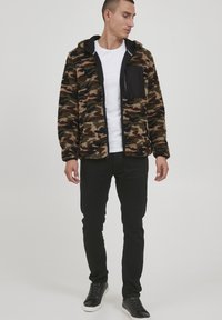 Camouflage fleece jacket with a hood and black chest pocket, worn over a white shirt. Paired with black pants and black sneakers.
