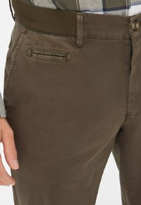 Pantaloni di cotone marroni con davanti piatto, tasche laterali e chiusura con zip. Dotati di una spessa vita elastica verde per comfort.