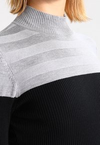 Gestrickter Pullover mit hohem, geripptem Kragen, mit einem gestreiften grauen oberen Abschnitt und einem einfarbigen schwarzen, gerippten unteren Abschnitt.