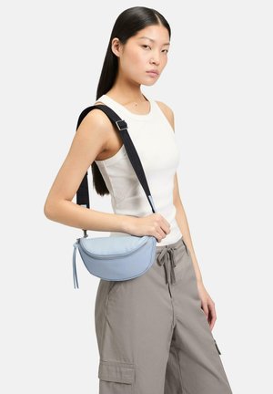 Hellblaues Halbmond-Umhängetäschchen mit Reißverschluss und einem strukturierten schwarzen Riemen, quer über den Körper getragen, kombiniert mit einem weißen Tanktop und grauen Hosen.