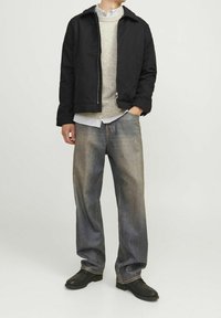 Giacca nera con zip frontale, maglione lavorato a maglia grigio chiaro, camicia a righe con bottoni, jeans larghi sbiaditi e scarpe nere.